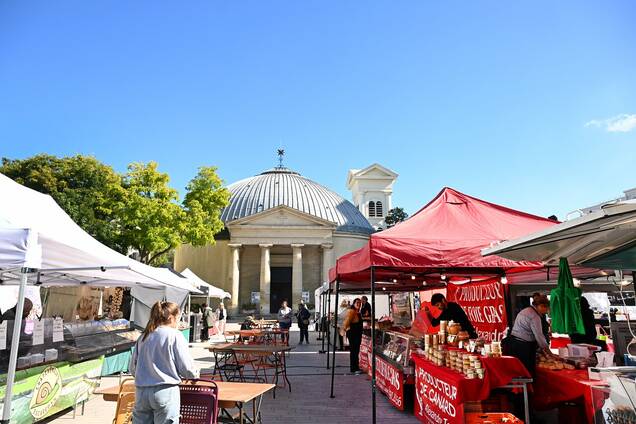 Photo des stands du marché aux producteurs devant l'église saint pierre saint paul en un jour ensoleillé avec du monde en train de manger sur les tables devant les stands et des gens qui font la queue.