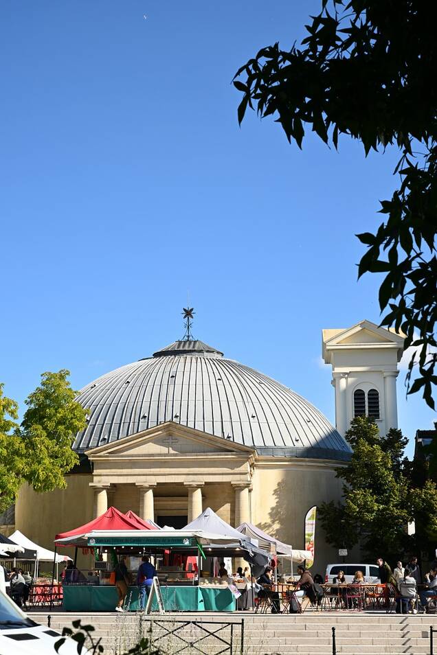 Photo des stands du marché aux producteurs devant l'église saint pierre saint paul en un jour ensoleillé avec du monde en train de manger sur les tables devant les stands et des gens qui font la queue.