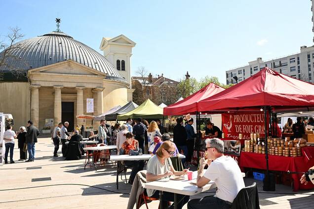 Photo des stands du marché aux producteurs devant l'église saint pierre saint paul en un jour ensoleillé avec du monde en train de manger sur les tables devant les stands et des gens qui font la queue.