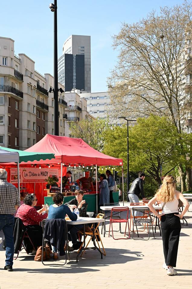 Photo des stands du marché aux producteurs devant l'église saint pierre saint paul en un jour ensoleillé avec du monde en train de manger sur les tables devant les stands et des gens qui font la queue.
